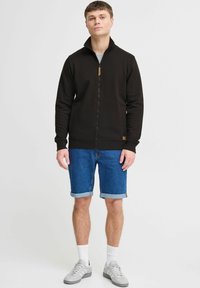 Black zip-up jacket with high collar, combined with blue denim shorts. Gray sneakers and white crew socks complete the outfit.