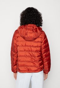 Person med lockigt hår bär en orange huva pufferjacka och ljusblå jeans, står vänd bort från kameran mot en enfärgad bakgrund.