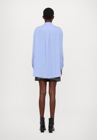 Camicia azzurra chiara a maniche lunghe con colletto rotondo, caratterizzata da una piega sul retro e polsini con bottoni. Indossata sopra una gonna nera a strati e stivaletti.