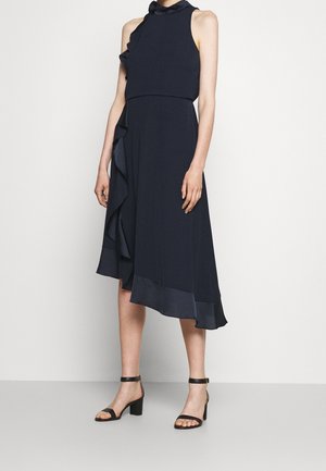 Femme portant une robe midi asymétrique sans manches bleu marine avec un détail en volants et des sandales à talons blocs noires à bride cheville.