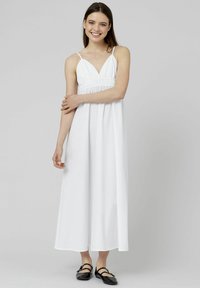 Robe maxi blanche avec bretelles spaghetti ajustables, une taille froncée et un décolleté en V. Tissu lisse et design longueur complète. Chaussures plates noires.