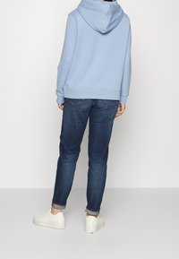 Lichtblauwe hoodie met een relaxte pasvorm, gecombineerd met donkerblauwe jeans en witte sneakers, wat een casual en comfortabele uitstraling vanaf de achterkant laat zien.