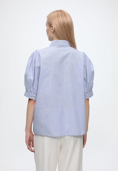 Femme blonde debout de dos, portant un chemisier bleu clair à manches courtes bouffantes et un pantalon blanc sur un fond uni.