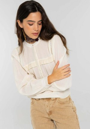 Femme aux longs cheveux bruns portant un chemisier blanc boutonné et un jean beige, écharpe autour du cou, regardant vers le bas avec les bras croisés.