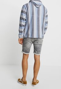 Camiseta con capucha a rayas grises y azules, combinada con pantalones cortos de mezclilla grises. Zapatos marrones con suelas blancas. Diseño casual, ajuste relajado.