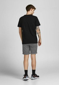 T-shirt en coton noir, shorts gris avec ourlets roulés, et chaussures de sport noires. Le modèle montre un tatouage sur le bras droit.