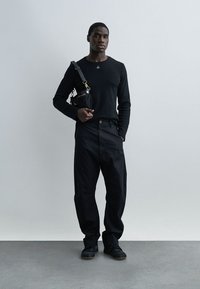 Άνδρας με ολόμαυρη εμφάνιση, φοράει μακρυμάνικο πουκάμισο, άνετο παντελόνι, μαύρα παπούτσια και μικρή μαύρη τσάντα ώμου, στέκεται μπροστά σε έναν απλό τοίχο.