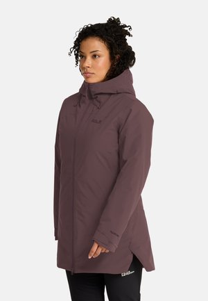 Donna con capelli ricci che indossa una lunga giacca viola con cappuccio, in piedi davanti a uno sfondo chiaro e semplice, rivolta verso sinistra.
