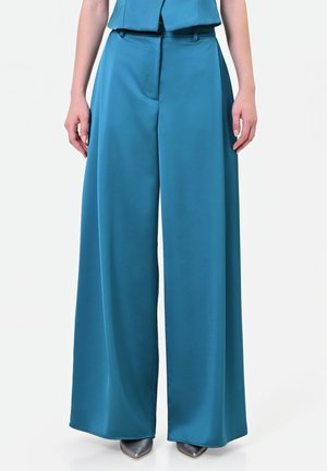 Pantaloni a gamba larga color turchese con cuciture frontali, indossati con un top turchese abbinato, abbinati a scarpe metalliche con punta a punta su sfondo bianco.