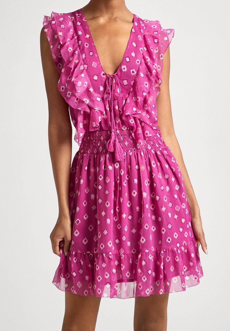 Robe rose sans manches avec motifs losanges blancs et rose foncé, épaules et ourlet à volants, profond décolleté en V avec cordons à nouer et taille élastique.