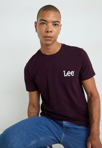 Bordeaux bomulds t-shirt med et hvidt "Lee" logo på venstre bryst, parret med blå denim jeans, siddende mod en lys baggrund.
