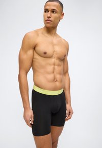 Μυώδης άνδρας που φοράει μαύρα μποξεράκια με κίτρινη ζώνη Calvin Klein, κοιτάζει προς τα αριστερά του σε απλό φωτεινό φόντο.