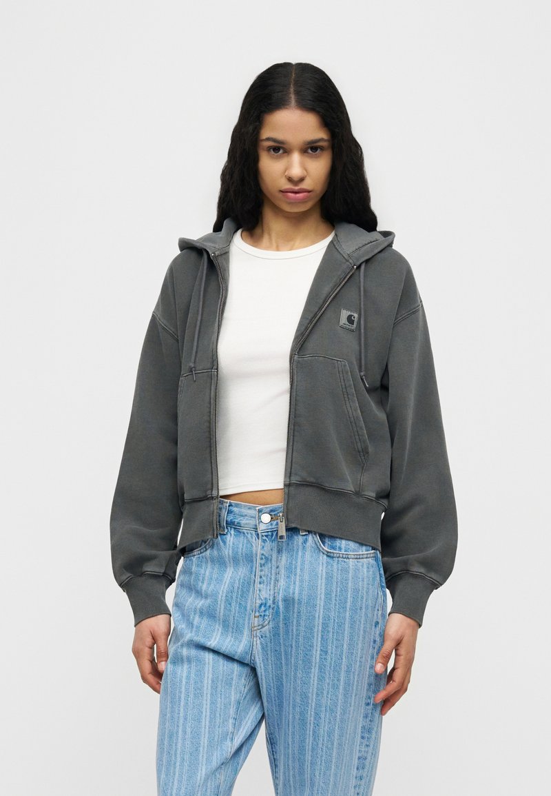 Giovane donna con lunghi capelli ricci che indossa una felpa grigia con zip, una maglietta bianca corta e jeans a righe blu, in piedi davanti a uno sfondo neutro.