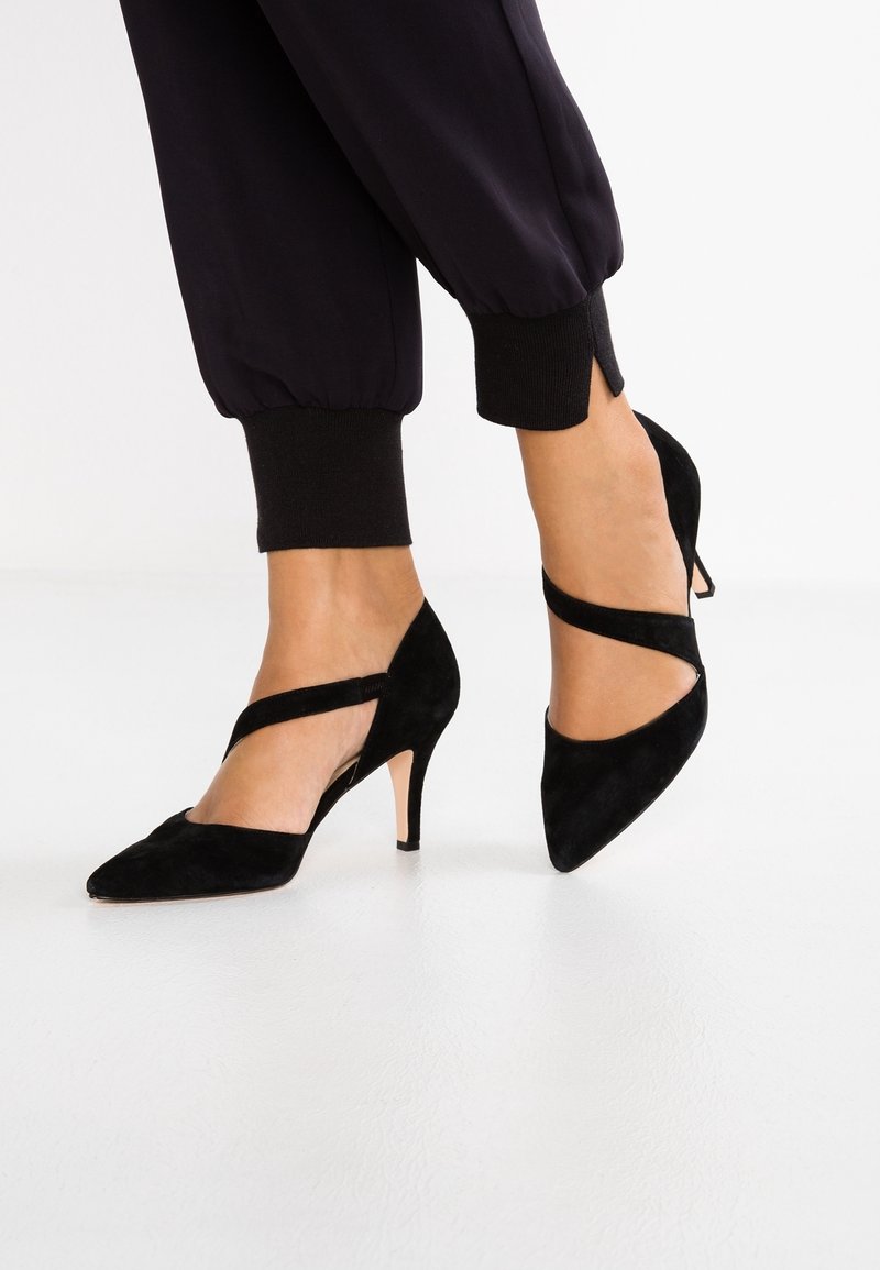 Pieds portant des escarpins en daim noir à bouts pointus avec des brides croisées, associés à un pantalon noir retroussé sur un fond blanc uni.