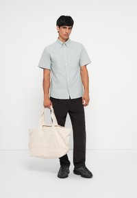 Light blue short-sleeve button-up shirt, dark pants, black sneakers, holding a cream-colored tote bag with dual handles. Simple design.