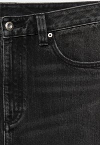 Gros plan sur un jean en denim noir montrant la ceinture, le bouton, la boucle de ceinture, le rivet et les détails de couture de la poche avant.