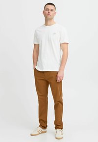 Bílá bavlněná trička s malou zlatou grafikou, spárovaná s úzkými hnědými kalhotami a bílými teniskami. Jednoduchý, volnočasový outfit.