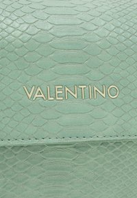 Mintgrön texturerad läderyta med gyllene metallbokstäver som stavar "VALENTINO" centrerat horisontellt.