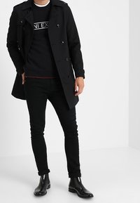 Trench-coat noir à double boutonnage, porté sur un pull noir avec un texte blanc. Jean noir et bottes noires brillantes complètent la tenue.