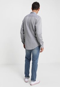 Camisa de manga larga gris claro con cuello abotonado, canesú trasero y dobladillo curvado, combinada con vaqueros azules y zapatillas blancas.