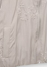 Giacca grigia chiara con intricati ricami floreali bianchi, chiusura frontale con zip e un tessuto morbido e texturizzato con orlo arricciato.