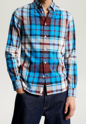 Mand iført blåt, rødt og hvidt ternet skjorte med krave, kombineret med mørkeblå jeans, hænder i lommerne, neutral baggrund.