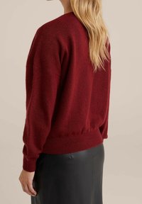 Burgundowa sweter z dzianiny z okrągłym dekoltem, obniżonymi ramionami, ściągaczami na rękawach i marszczonym dołem, noszony na czarnej skórzanej spódnicy.