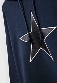 Sweat à capuche bleu marine avec un grand logo étoile noire bordé de blanc, présentant des accents en tissu texturé et des cordons à pompons.