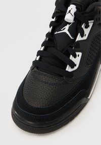 Κοντινό πλάνο μαύρου αθλητικού παπουτσιού Air Jordan με υφή δέρματος, μαύρα κορδόνια, λευκές πλαστικές οπές και λευκό λογότυπο Jumpman στη γλώσσα και στο κλείδωμα των κορδονιών.