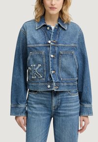 Przycięta kurtka dżinsowa w jasnoniebieskim kolorze z metalowymi guzikami, dwoma przednimi kieszeniami oraz ozdobną naszywką w kształcie litery 'K' na lewej kieszeni.