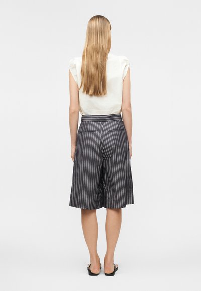 Γυναίκα με μακριά ξανθά μαλλιά, ντυμένη με λευκό αμάνικο τοπ και μαύρο σορτς με ριγέ σχέδιο, ψηλόμεσο και με φαρδιά μπατζάκια, στέκεται γυρισμένη προς τα πίσω.