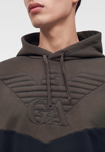 Jeune personne portant un sweat à capuche marron et noir avec un logo aigle brodé et les lettres GA sur la poitrine, gros plan sur le cou et le bas du visage.