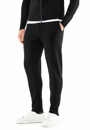 Pantalons noirs ajustés avec poches latérales, en tissu texturé et coupe slim, accompagnés de baskets blanches pour le contraste. Design minimaliste.