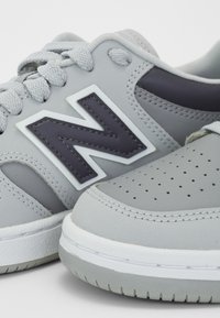 Gros plan de chaussures de sport grises avec un bout perforé, des lacets texturés et une grande lettre noire N sur le côté.