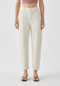 Pantalones de tiro alto color crema fabricados en algodón, con bolsillos laterales, piernas rectas y detalles de costura sutiles. Combinados con sandalias negras.
