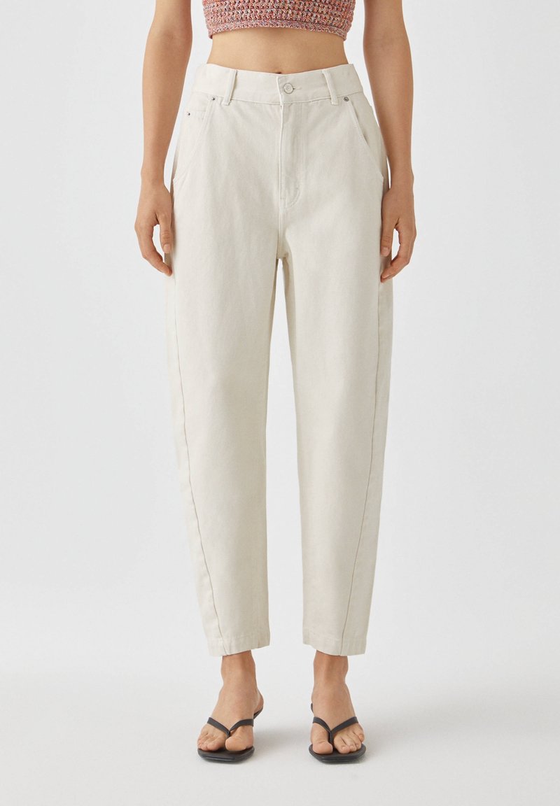 Pantalones de tiro alto color crema fabricados en algodón, con bolsillos laterales, piernas rectas y detalles de costura sutiles. Combinados con sandalias negras.