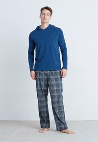 Marineblauwe hoodie met lange mouwen en een voorzak, gecombineerd met grijs geruite pyjamabroek. Zachte stof met een ontspannen pasvorm. Blote voeten.