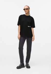 Sort oversized t-shirt med broderet logo, parret med mørke jeans og kraftige sorte støvler, sat mod en simpel hvid baggrund.