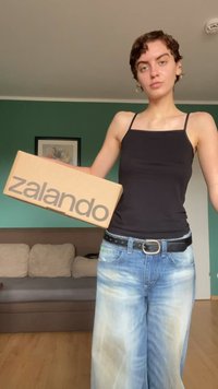 Cardboard box featuring "zalando" logo in gray. Individual wearing a black tank top and light blue wide-leg jeans, holding the box.