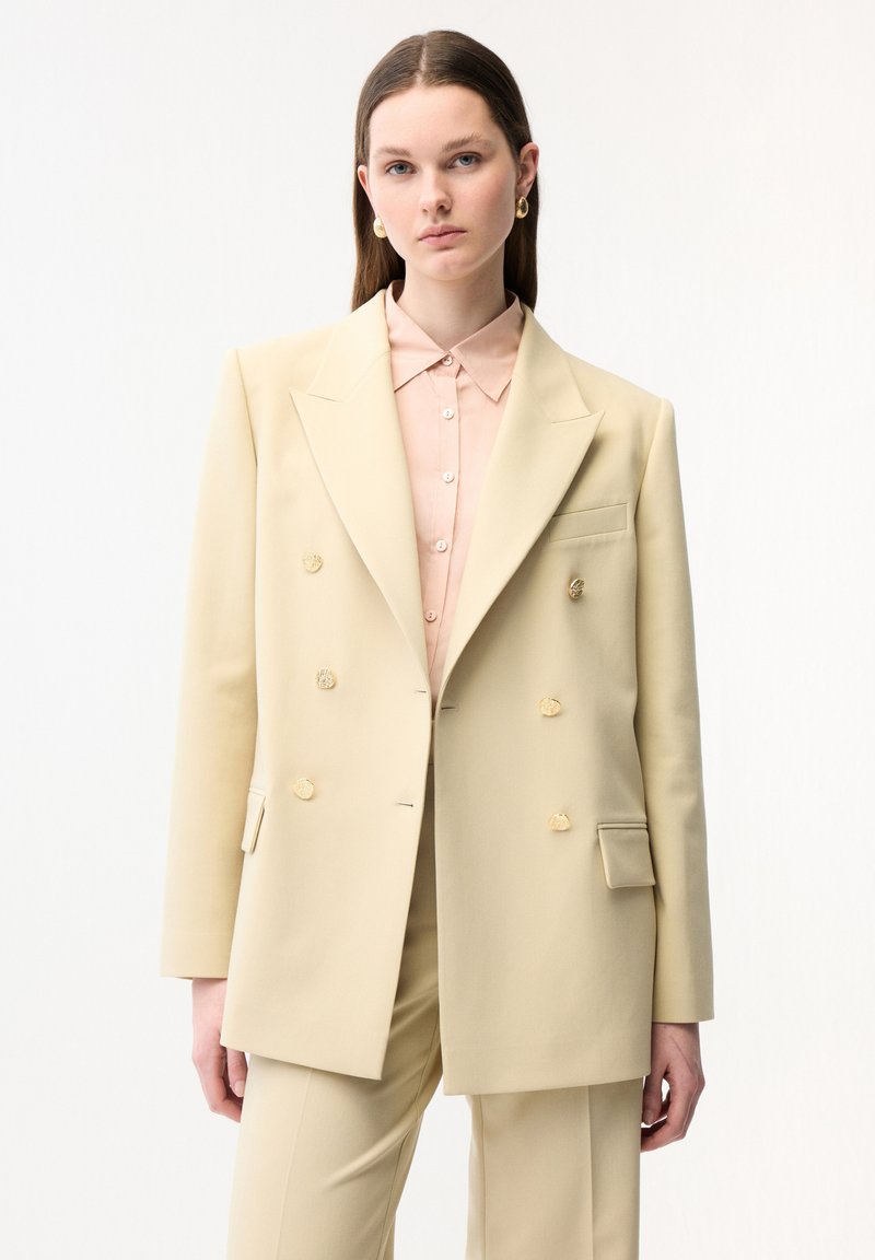 Joven mujer que lleva un blazer beige de doble botonadura y pantalones a juego, con una camisa de botones color rosa pálido y pendientes de aro dorados.