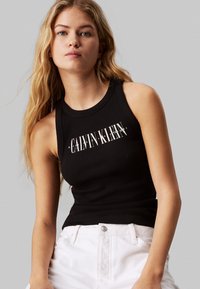 Top a canotta nero a coste con "CALVIN KLEIN" stampato in bianco sul petto. Indossato con pantaloni bianchi, con dettagli sui bottoni.