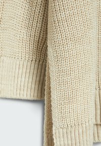 Beige gebreide trui met geribbelde manchetten en onderrand, die een stevige textuur en een iets losse weving laat zien, benadrukt warmte en comfort.