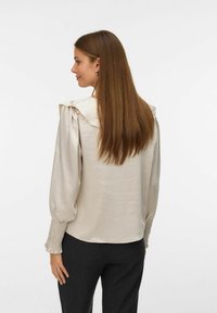Jasno-beżowa bluzka z falbaniastym kołnierzem, bufiastymi rękawami i teksturowanymi mankietami. Noszona z ciemnymi spodniami, prezentując prosty, elegancki design.
