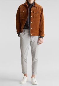 Ruskea mokkatakki, jossa kaksi rintataskua, yhdistettynä tummakuviolliseen paitaan ja vaaleanharmaisiin ruutuhousuihin sekä valkoisiin tennareihin.