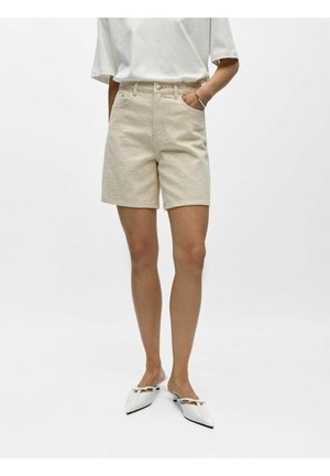 Femme portant un short beige taille haute à motif subtil, un haut blanc à manches courtes, un bracelet argenté et des chaussures blanches à enfiler à bout pointu.