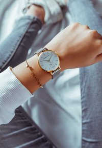 Hopeinen verkko-ristikko kellossa on valkoinen taulu ja kultaiset yksityiskohdat, jotka sisältävät tuntimerkit ja pienen päivämääränäytön. Mukana tulee herkkä rannekoru.