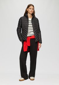 Schwarze gesteppte Jacke mit hohe Kragen, getragen über einem weiß gestreiften Hemd. Roter Pullover um die Taille gebunden, schwarze Hose und helle Sneaker.