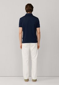 Hombre de pie de espaldas vistiendo una camiseta tipo polo azul marino, pantalones blancos y alpargatas verdes, sobre un fondo gris liso.