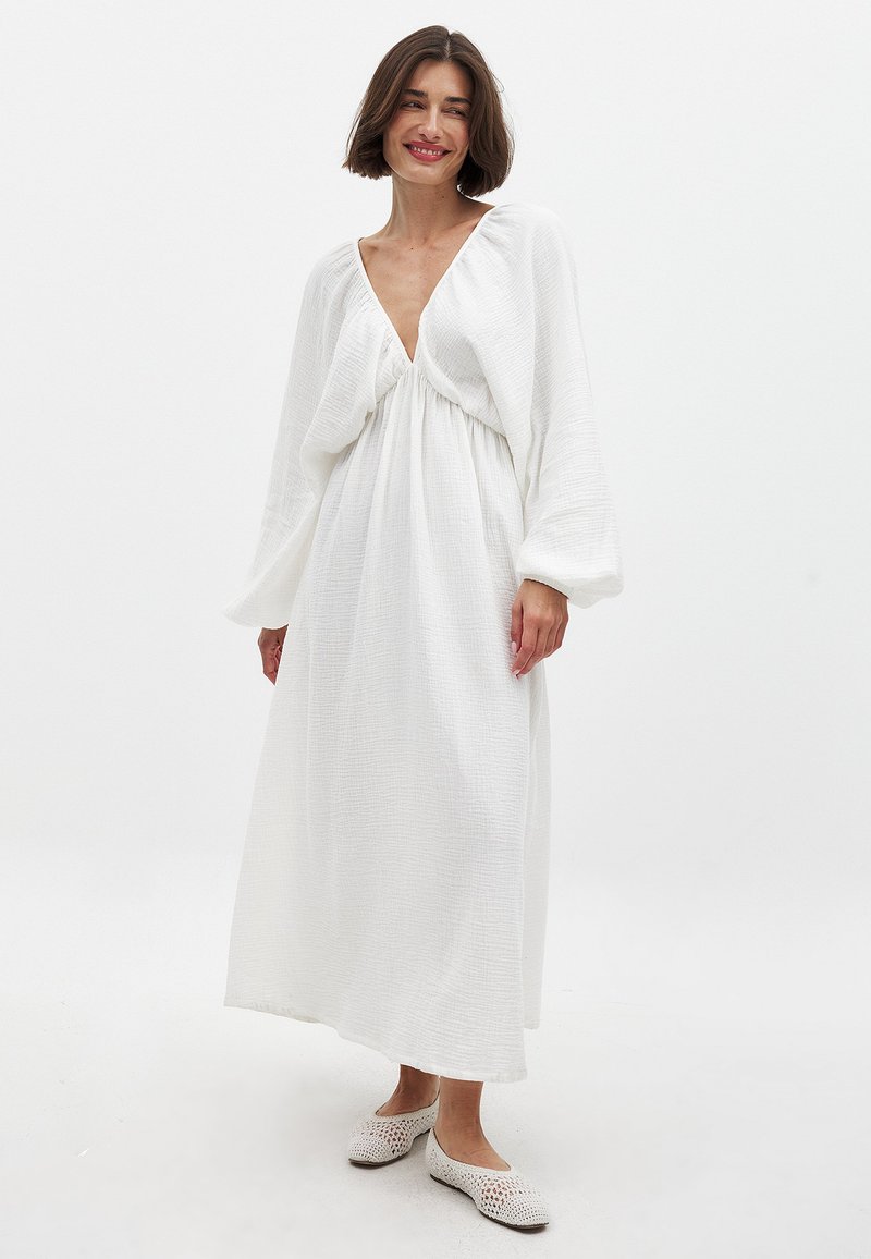 Langes weißes Kleid aus strukturiertem Stoff, mit tiefem V-Ausschnitt und weiten Ärmeln. Getragen mit leichten gehäkelten Ballerinas.