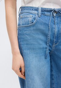 Κοντινή λήψη ενός ατόμου που φοράει τζιν παντελόνι ανοιχτού μπλε χρώματος με υφασμάτινη ζώνη δεμένη στη μέση, συνδυασμένο με λευκό πουκάμισο.